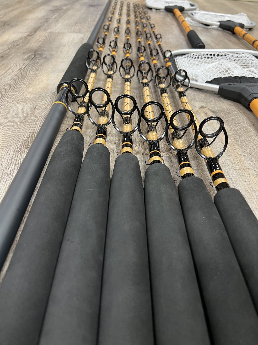 A row of offshore rods with gold and black decorative wraps, oversized guides and foam grips, on a gray wood floor